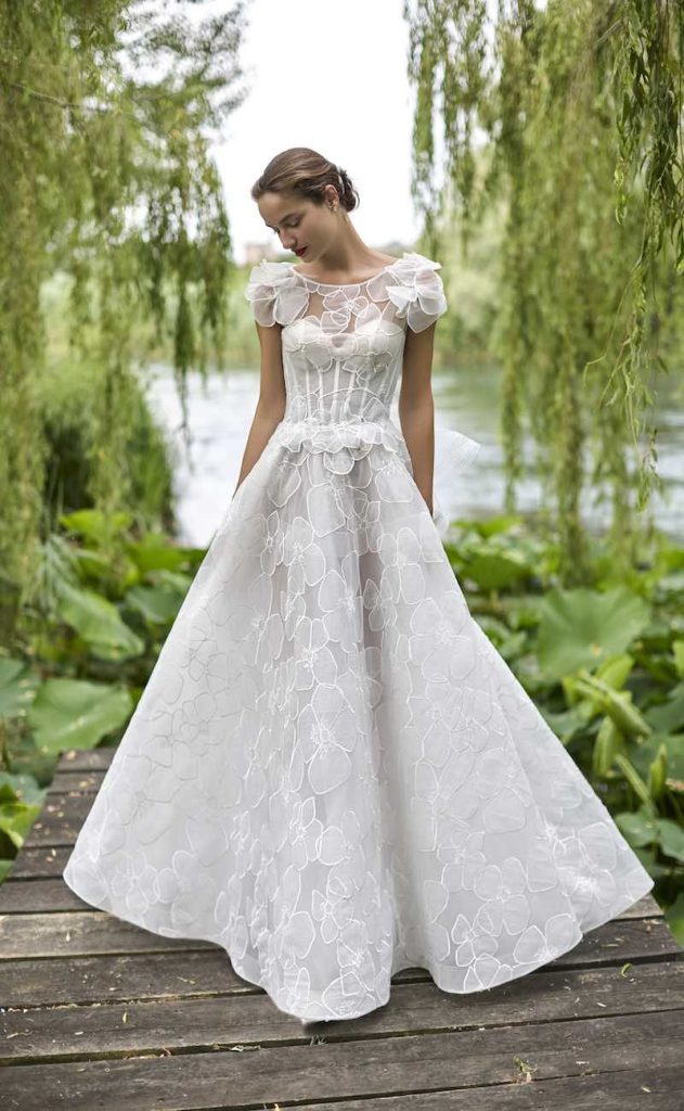 In questa foto, una modella posa con un abito da sposa a A in tulle con disegni floreali e corpetto steccato della collezione sposa Elisabetta Polignano 2026