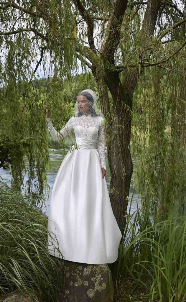 In questa foto, una modella posa su un albero con un abito da sposa ad A con gonna in mikado e corpetto a maniche lunghe in pizzo della collezione sposa Elisabetta Polignano 2026