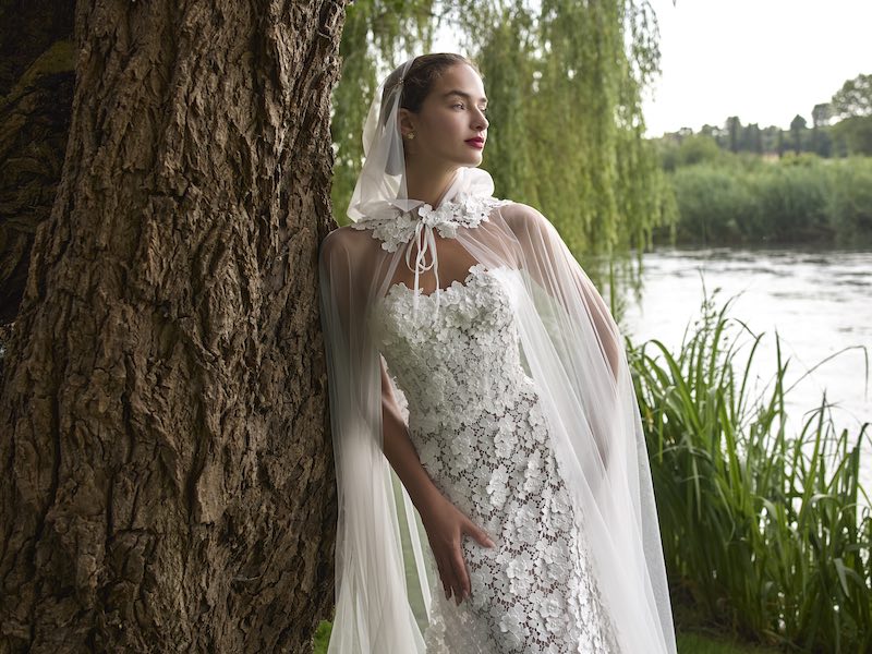 In questa foto, una modella posa con un abito da sposa in pizzo e una cappa della collezione sposa Elisabetta Polignano 2026