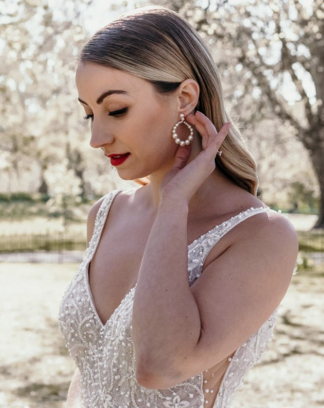 In questa foto, una sposa in abito elegante si tocca l’orecchio per sistemare un orecchino a cerchio con perle. Ha i capelli lisci e un trucco raffinato con rossetto rosso. L’immagine mostra un look sposa classico e glamour, offrendo un esempio concreto di come scegliere trucco sposa per un matrimonio dal gusto sofisticato.