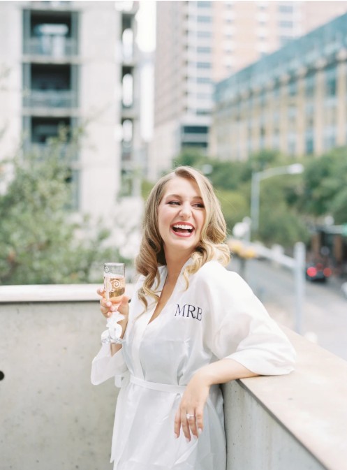 In questa foto, una futura sposa bionda sorride raggiante mentre si trova su una terrazza urbana. Indossa un accappatoio bianco personalizzato con le sue iniziali e tiene in mano un flute di champagne. I suoi capelli sono raccolti in morbide onde e il trucco è fresco, elegante, con labbra rosate e sguardo definito. Un'immagine che suggerisce come scegliere trucco sposa per un look radioso e sofisticato.