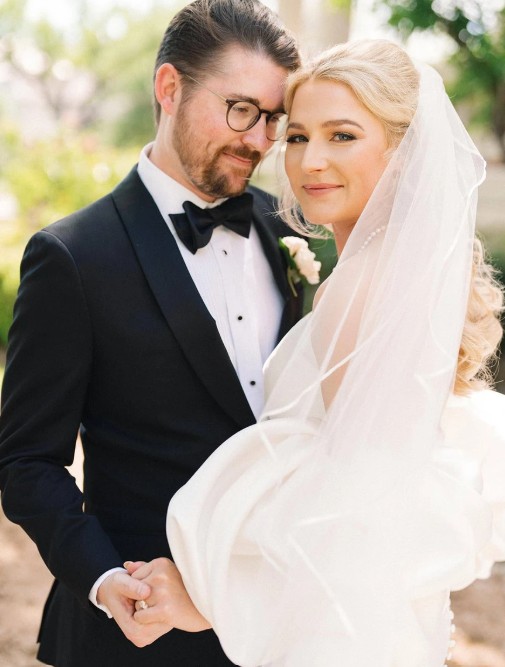 In questa foto una coppia di sposi è ritratta in un tenero abbraccio all’aperto. Lei, con un velo leggero che le avvolge le spalle, sorride guardando l’obiettivo; lui indossa uno smoking nero con papillon e occhiali, e la guarda con dolcezza. La luce naturale valorizza il make-up luminoso e raffinato della sposa, perfetto per chi vuole capire come scegliere trucco sposa per un matrimonio di giorno.