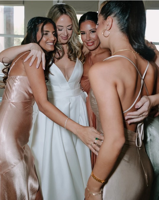 In questa foto una sposa in abito bianco con scollo a V viene abbracciata affettuosamente dalle sue damigelle, che indossano abiti satinati nei toni nude e champagne. Il gruppo è ritratto in un momento di complicità e felicità. Il trucco fresco e naturale della sposa si integra perfettamente con l’atmosfera intima, offrendo spunti su come scegliere trucco sposa che valorizzi la propria bellezza senza eccessi.