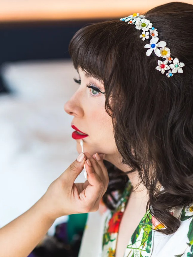 In questa foto, una donna con capelli castani e frangia riceve un ritocco di rossetto rosso, indossando un cerchietto con fiori colorati e un kimono floreale. Il suo trucco deciso e vintage mostra come scegliere trucco sposa possa significare esaltare la personalità e valorizzare uno stile unico e creativo.
