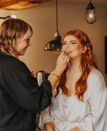 In questa foto, una make-up artist sta applicando il rossetto a una sposa dai lunghi capelli rossi, sorridente e rilassata. L’atmosfera è calda e intima, con luci soffuse e dettagli rustici. L'immagine suggerisce come scegliere trucco sposa in base all'incarnato e alla palette cromatica del matrimonio, puntando su tonalità calde e naturali.