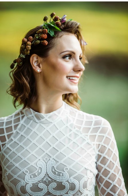 In questa foto, una sposa sorride guardando verso destra, con una corona floreale fatta di more e foglie tra i capelli mossi. Indossa un abito bianco a collo alto con motivo a rete. L’immagine esprime naturalezza e freschezza, ed è perfetta per ispirarsi su come scegliere trucco sposa in stile boho o campestre.