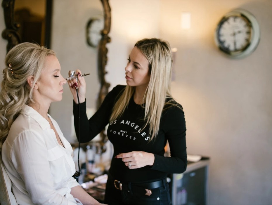 In questa foto, una truccatrice professionista applica il make-up a una sposa utilizzando l’aerografo, mentre la futura sposa siede con gli occhi chiusi e un'espressione rilassata. La scena si svolge in una stanza elegante, con specchi decorati e luci soffuse. L’immagine è un perfetto esempio di come scegliere trucco sposa puntando su tecniche professionali e un risultato naturale ma impeccabile.
