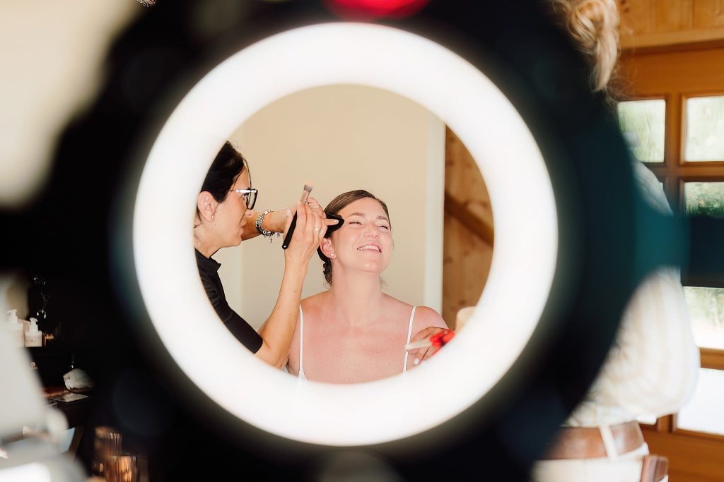 In questa foto, vista attraverso un anello di luce, una truccatrice sta applicando il make-up a una sposa che sorride a occhi chiusi. Il momento è intimo e rilassato, catturando la complicità del prepararsi. Questa immagine rappresenta un esempio autentico su come scegliere trucco sposa che faccia sentire bene nel proprio giorno speciale.