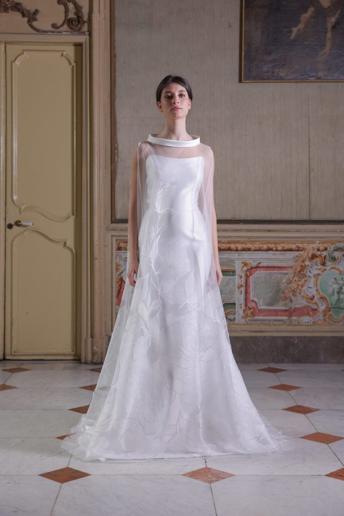 In questa foto, una modella indossa un abito da sposa della collezione Essenza Atelier Benedetta 2026 modello a sirena con cappa jacquard 