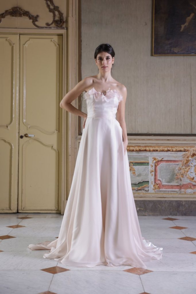In questa foto, una modella indossa un abito da sposa della collezione Essenza Atelier Benedetta 2026, colore rosa cipria, modello ad A con scollo a cuore e fiori 3D sullo scollo
