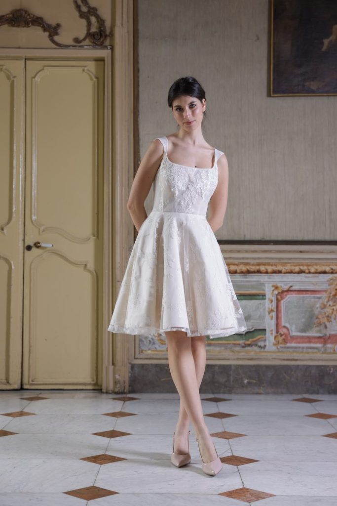 In questa foto, una modella indossa un abito da sposa della collezione Essenza Atelier Benedetta 2026 modello corto con gonna a corolla e scollo quadrato 