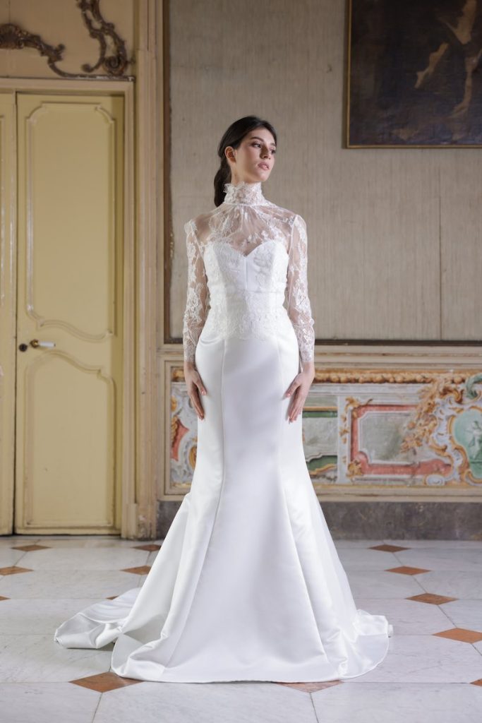 In questa foto, una modella indossa un abito da sposa della collezione Essenza Atelier Benedetta 2026 modello semisirena in mikado con corpetto a collo alto ricamato in pizzo 