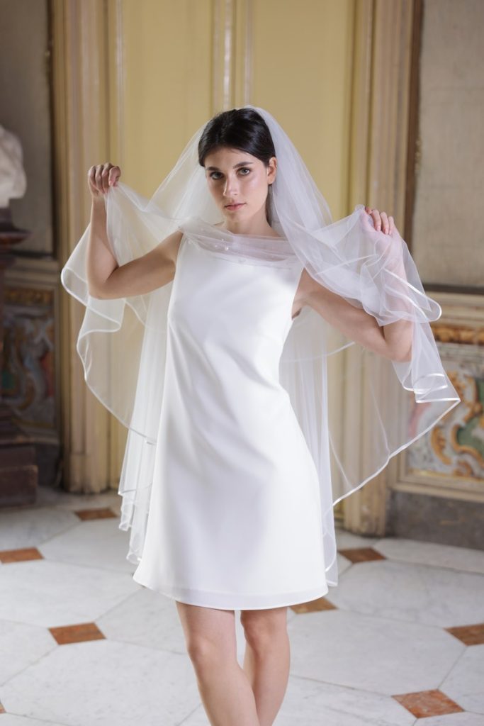 In questa foto, una modella indossa un abito da sposa della collezione Essenza Atelier Benedetta 2026 modello a tubino con velo corto in tulle abbinato