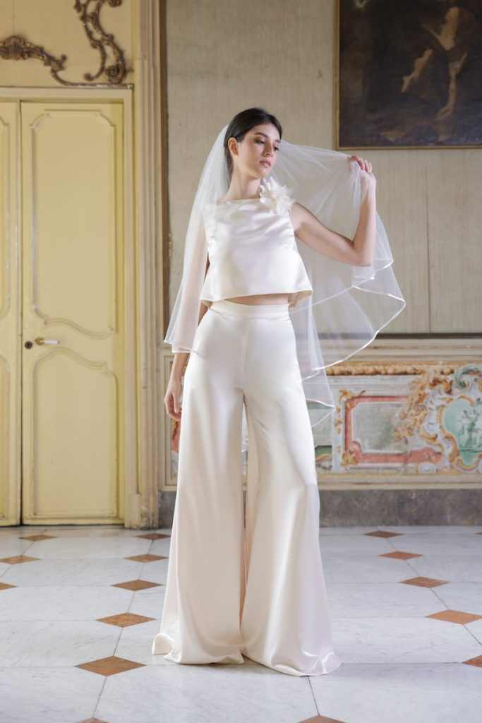 In questa foto, una modella indossa un abito da sposa della collezione Essenza Atelier Benedetta 2026, colore champagne, modello pantaloni palazzo con crop top decorato con un fiore 3D sulla spalla