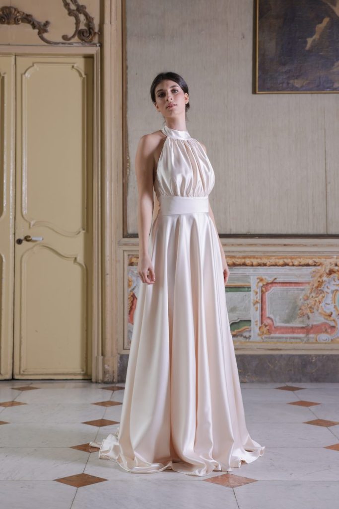 In questa foto, una modella indossa un abito da sposa della collezione Essenza Atelier Benedetta 2026, colore rosa cipria, modello scivolato con scollo all'americana