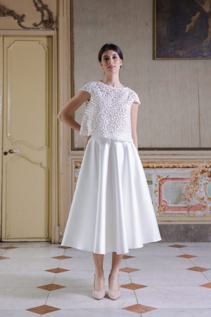 In questa foto, una modella indossa un abito da sposa della collezione Essenza Atelier Benedetta 2026 modello due pezzi con gonna a corolla e canotta ricamata