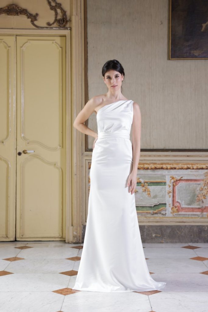 In questa foto, una modella indossa un abito da sposa della collezione Essenza Atelier Benedetta 2026 modello a sirena, in tessuto mikado, con scollo monospalla