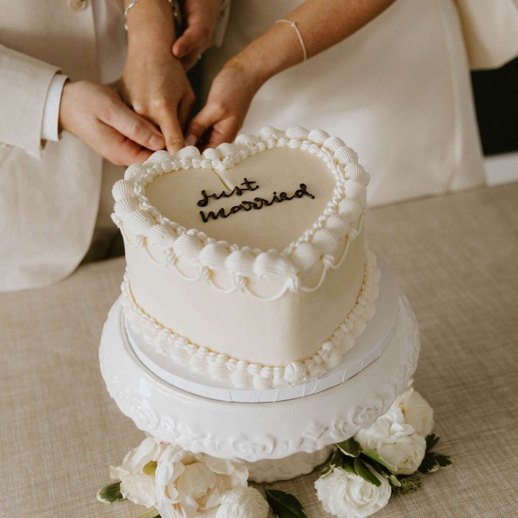 In questa foto che mostra Quale torta scegliere per il matrimonio" si vede una torta nuziale a forma di cuore, di colore bianco, decorata con glassa ondulata lungo i bordi e con la scritta "just married" in cima, realizzata con cioccolato scuro. La torta è posizionata su un'alzata bianca in ceramica decorata, circondata da fiori bianchi alla base. Due persone stanno tagliando insieme la torta, tenendosi per mano in un gesto affettuoso.