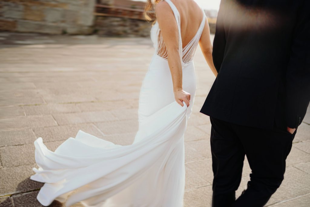 In questa foto si vede uno scatto di Ilaria&Andrea durante una passeggiata mano nella mano. Lei indossa un abito da sposa di colore bianco, aderente e con schiena scoperta decorata da fili sottili. La scena è luminosa e il vento muove dolcemente la gonna dell’abito, mentre lui, di spalle, indossa un completo di colore nero.