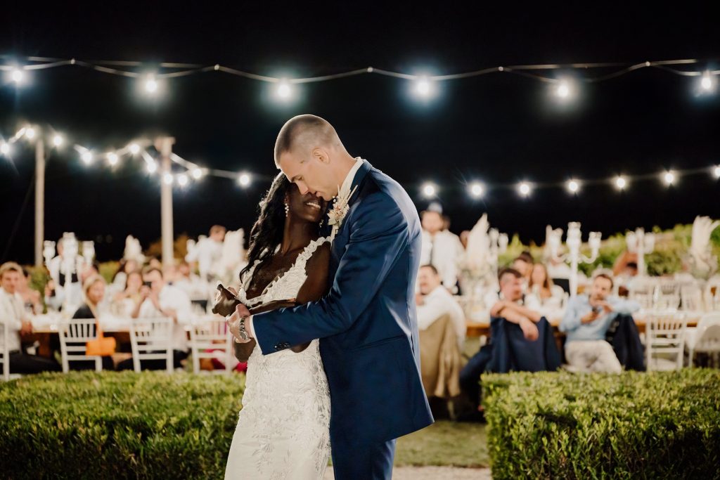 In questa foto si vede uno scatto di Ilaria&Andrea durante il primo ballo di una giovane coppia. I due sposi si abbracciano sotto un cielo notturno, illuminati da una fila di luci sospese. Lei indossa un abito da sposa di colore bianco con decorazioni in pizzo, lui un completo elegante di colore blu. Intorno a loro, gli invitati assistono commossi alla scena.