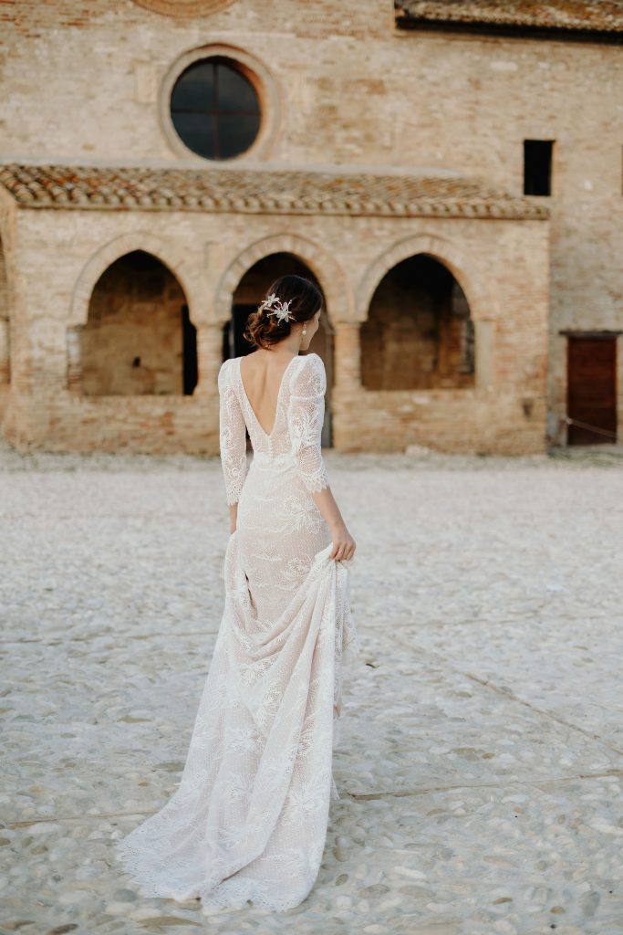 In questa foto si vede uno scatto di Ilaria&Andrea durante un servizio fotografico di nozze: la sposa, ripresa di spalle, cammina su un ampio cortile in pietra davanti a una chiesa antica in pietra di colore beige. Indossa un abito da sposa lungo e aderente, di colore bianco panna con motivi in pizzo e maniche lunghe. I capelli sono raccolti in uno chignon basso decorato con un fermaglio floreale chiaro.