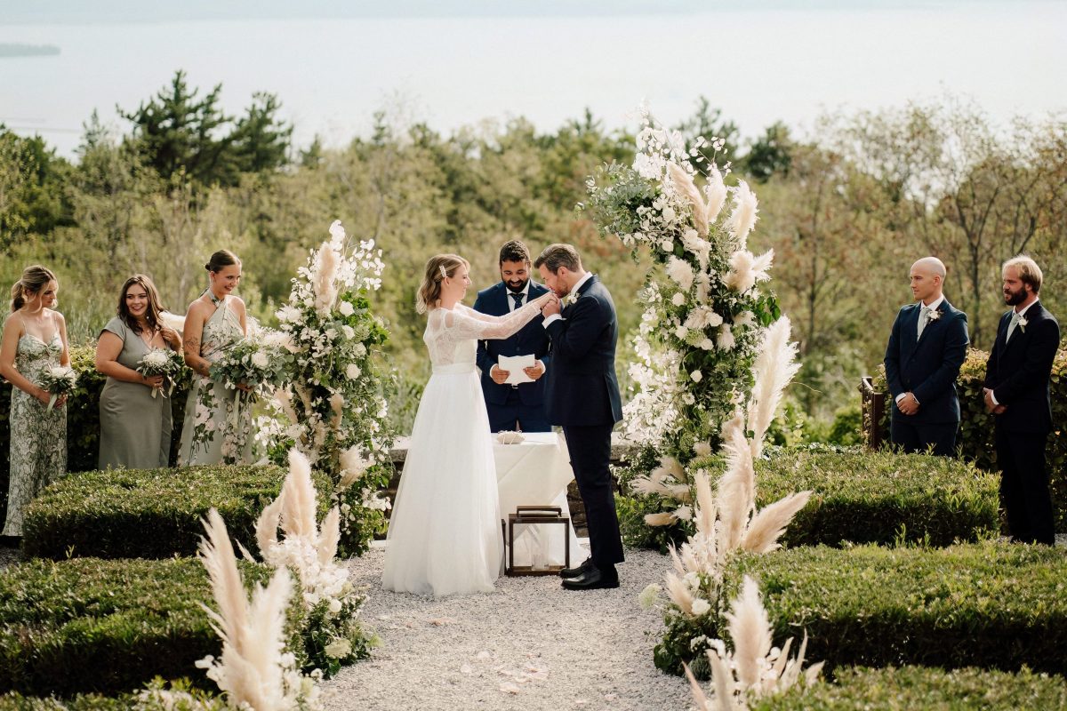 In questa foto si vede uno scatto di Ilaria&Andrea durante la cerimonia all’aperto. I due sposi sono al centro della scena, circondati da composizioni floreali bianche e pampas. La sposa indossa un abito lungo di colore bianco e tiene per mano lo sposo, che indossa un completo elegante di colore blu scuro. Sullo sfondo si intravede il paesaggio con alberi e un lago in lontananza