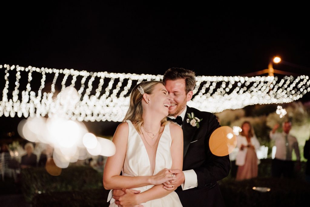 In questa foto si vede uno scatto di Ilaria&Andrea durante il ricevimento. Gli sposi sono avvolti da luci decorative sospese di colore bianco che brillano nella notte. I due sposi, entrambi sorridenti, sono stretti in un abbraccio tenero: lei indossa un abito da sposa con scollo profondo di colore bianco, lui un elegante completo da cerimonia di colore nero con camicia di colore bianco. Sullo sfondo, si intravedono alcuni invitati che tengono in mano piccole scintille luminose.