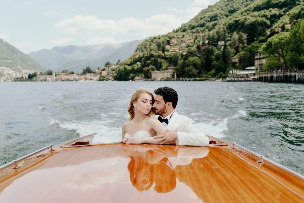 In questa foto si vede uno scatto di Ilaria&Andrea durante una romantica gita in barca sul lago. I due sposi sono in piedi su una barca in legno lucido, con lei che indossa un abito da sposa senza spalline di colore bianco, mentre lui, di fianco a lei, indossa uno smoking con giacca di colore bianco e papillon nero. Sullo sfondo si intravedono le colline verdi e le case affacciate sull’acqua, in un’atmosfera intima e suggestiva