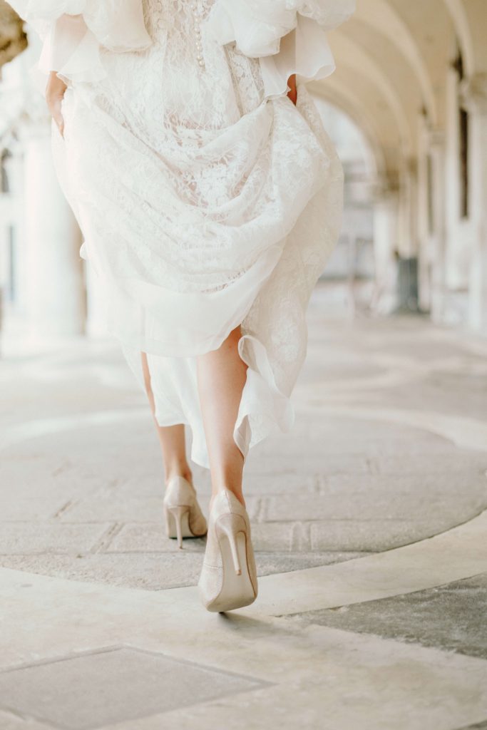 In questa foto si vede uno scatto di Ilaria&Andrea durante un servizio fotografico di nozze: un dettaglio in movimento mostra le gambe della sposa mentre cammina, sollevando leggermente l’abito di pizzo di colore bianco. Indossa scarpe con tacco alto di colore beige chiaro, camminando sotto un porticato elegante con archi e pavimento in pietra.