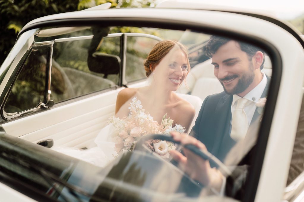 In questa foto si vede uno scatto di Ilaria&Andrea durante matrimonio nel momento in auto dopo la cerimonia. I due sposi sono seduti su una macchina d’epoca decappottabile di colore bianco. La sposa sorride e tiene un bouquet di fiori dai toni pastello, mentre lo sposo, che indossa un abito di colore nero con cravatta panna, sorride