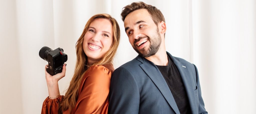 In questa foto si vede uno scatto di Ilaria&Andrea durante un momento divertente in studio. I due sono in piedi schiena contro schiena, entrambi sorridenti e con in mano una macchina fotografica. Ilaria indossa un abito lungo plissettato di colore rame, mentre Andrea ha un completo elegante di colore blu scuro con t-shirt di colore nero. Lo sfondo è una tenda di colore bianco che rende l’immagine luminosa ed essenziale.