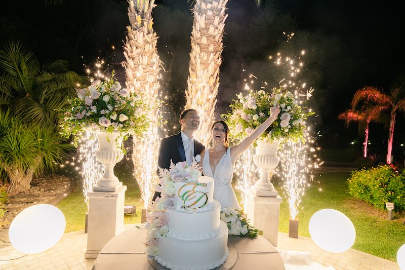 In questa foto, due sposi al taglio della torta nuziale a tre piani decoarata con le iniziali E e D e con fiori di colore rosa e bianco. Dietro di loro un'esplosione di fuochi d'artificio
