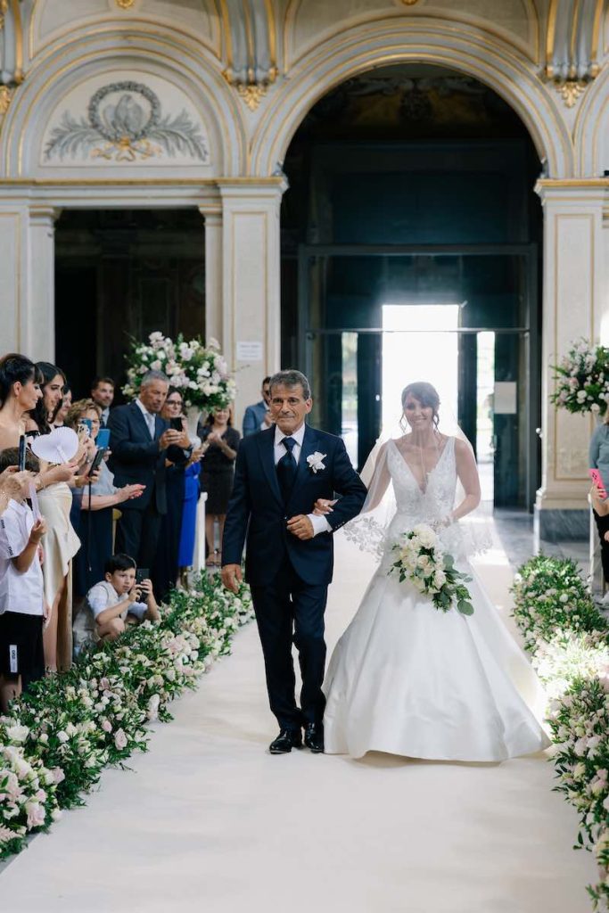 In questa foto, la sposa Eleonora entra nella Basilica di Santa Cecilia in Trastevere, emozionata con il papà tenendo nella mano destra un bouquet a cascata con fiori di colore bianco e attraversando un corridoio di fiori