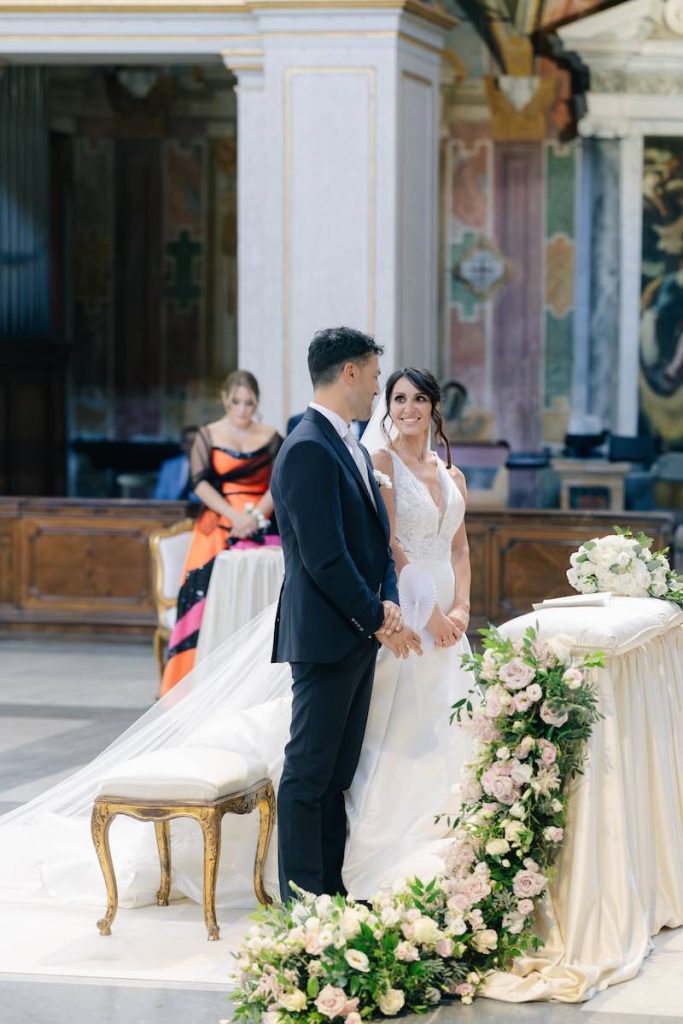 In questa foto, un momento della cerimonia religiosa di Eleonora e Danilo: i due sposi si guardano sorridenti all'altare. In primo piano, un cordone floreale di rose e fiori di colore bianco e rosa sviluppato da Giorgia Pirro