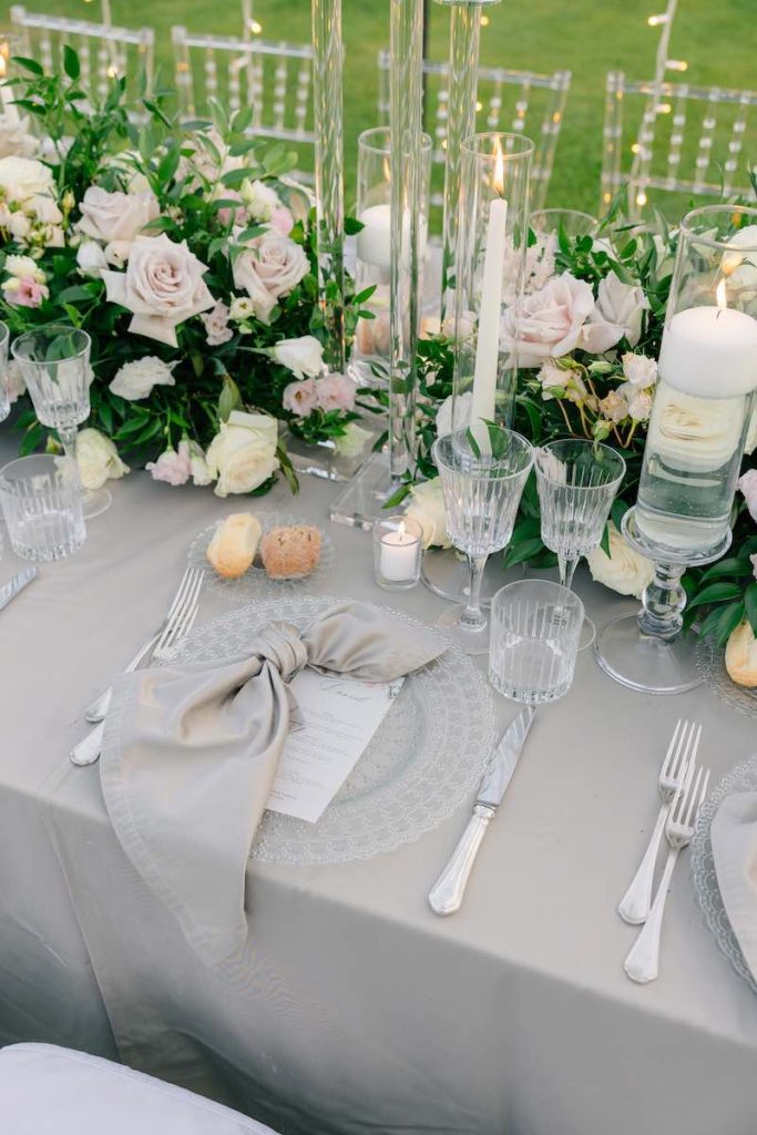In questa foto, una mise en place di matrimonio firmata da Giorgia Pirro realizzata con un tovagliato di colore grigio perla, con fiori di colore rosa e bianco, candele, candelabri, piatti in vetro trasparente e posate d'argento