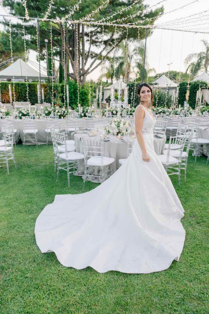 In questa foto, una sposa con un abito ampio con lungo strascico posa in giardino davanti all'allestimento del ricevimento di nozze nei toni del grigio perla, del rosa e del bianco con tavoli decorati con fiori, candele e candelabri