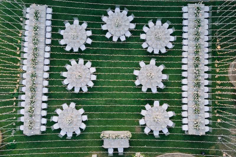 In questa foto, una vista dall'alto di un ricevimento in giardino con un mix di tavoli rotondi e imperiali sovrastati da un tetto di luci 