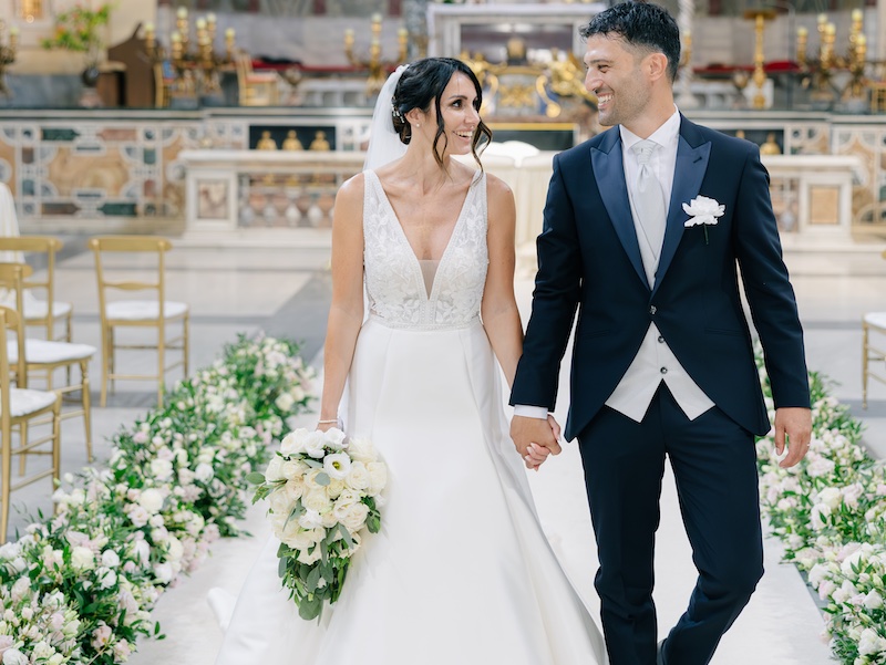In questa foto, Eleonora e Danilo escono dalla chiesa guardandosi negli occhi. I due sposi attraversano un coridoio di fiori di colore bianco progettato dalla Wedding Planner Giorgia Pirro