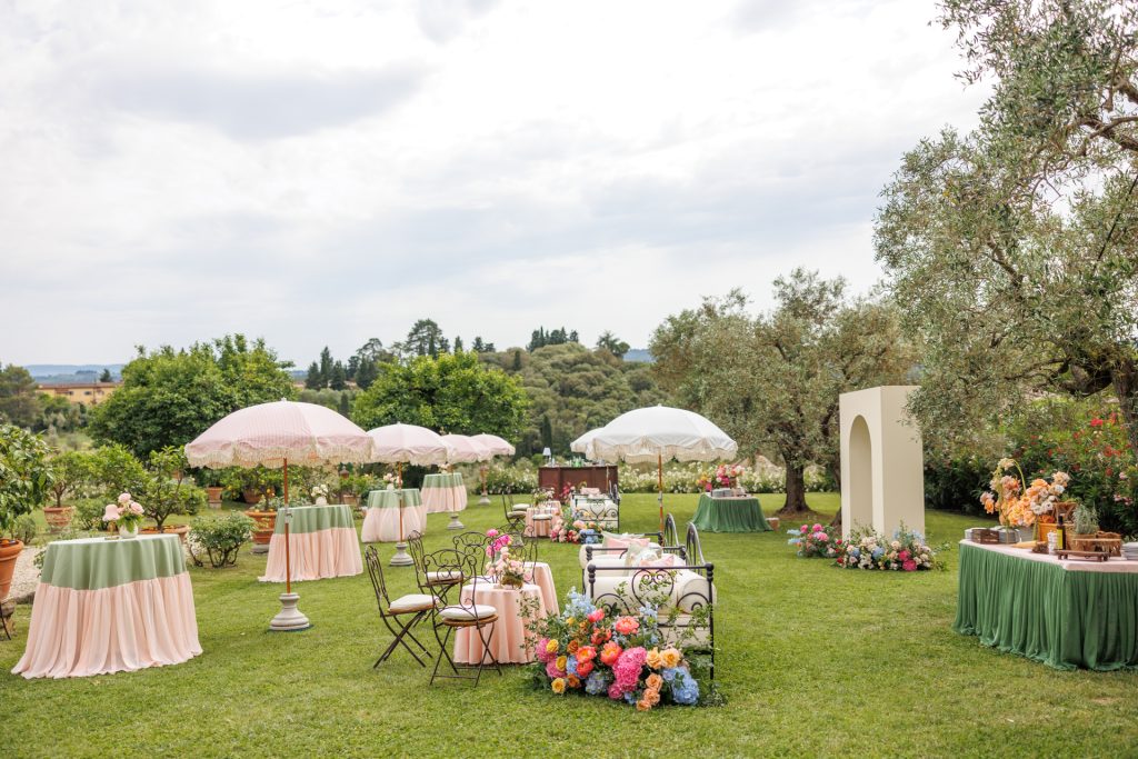 in questa foto del matrimonio di Emily e Mark firmato ester chianelli si vede Un grande prato verde ospita un raffinato allestimento per un aperitivo all’aperto, circondato da ulivi e alberi da giardino. Tavoli rotondi alti e bassi, rivestiti con tovaglie rosa cipria e verde salvia, sono distribuiti con armonia. Ogni tavolo è decorato con fiori freschi e piccole composizioni floreali. Numerosi ombrelloni dalle tinte pastello (rosa e bianco) con bordature in pizzo offrono ombra, creando un’atmosfera romantica e vintage. Sedute in ferro battuto bianco con cuscini chiari si affiancano a divanetti decorati con bouquet di fiori colorati (rose, ortensie, peonie in tonalità pesca, fucsia, azzurro, arancio). Sulla destra si trova un tavolo buffet con tovaglia verde, impreziosito da vasi e bottiglie decorative. Sullo sfondo si intravedono altre postazioni e un arco decorativo color panna.