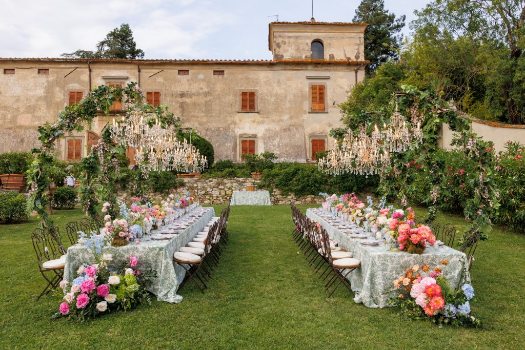 in questa foto del matrimonio di Emily e Mark firmato ester chianelli si vede un’elegante scena allestita per una cena all’aperto nel giardino di una villa dallo stile rustico. Al centro del prato, due lunghi tavoli rettangolari sono disposti parallelamente, coperti da tovaglie colore verde salvia con un delicato motivo floreale bianco. Attorno ai tavoli ci sono sedie in ferro battuto con cuscini bianchi.

Ogni tavolo è decorato con sontuose composizioni floreali dai colori vivaci: rose, peonie, ortensie e altri fiori in tonalità rosa, pesca, blu, lilla e bianco, disposte sia al centro che ai piedi dei tavoli.

Sopra i tavoli si sviluppano tre grandi archi ricoperti di foglie verdi e grappoli di fiori, da cui pendono maestosi lampadari di cristallo illuminati, che aggiungono un tocco lussuoso alla scena.

Sul fondo, si vede la facciata vissuta ma affascinante di una villa antica con persiane in legno e pareti scrostate, incorniciata da cespugli, fiori e un muro in pietra. L’atmosfera è serena, romantica e raffinata.