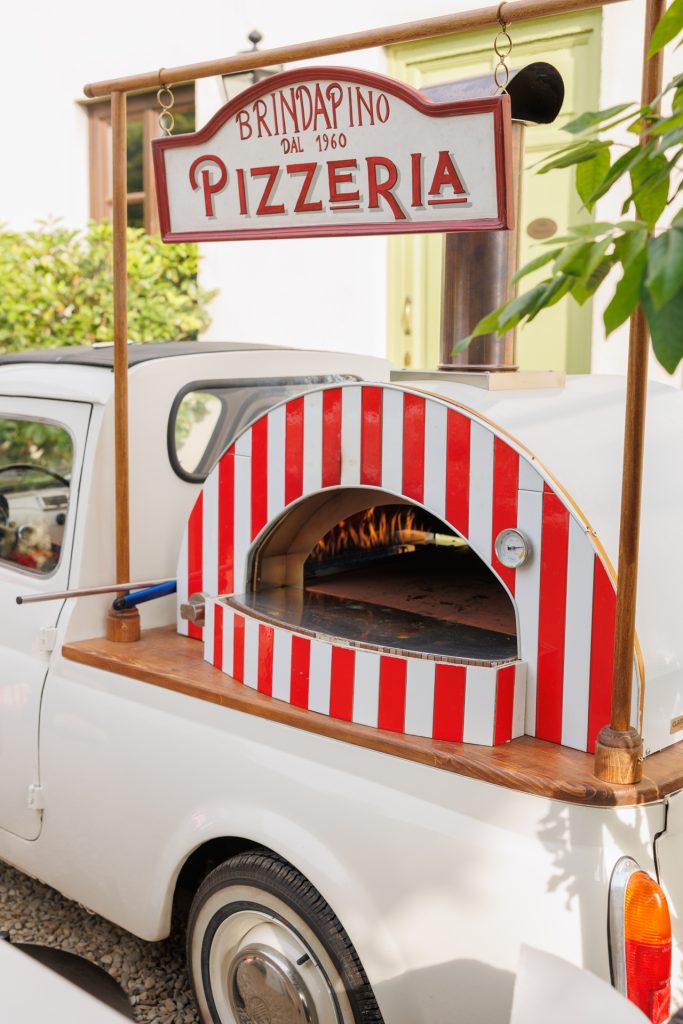 in questa foto del matrimonio di Emily e Mark firmato ester chianelli si vede Forno per pizza su una Fiat 500 vintage. Un piccolo forno a legna per pizza con decorazioni a righe rosse e bianche è montato sul retro di una Fiat 500 bianca. Sopra il forno, un’insegna recita "Pizzeria Brindapino dal 1960". Si vede il fuoco acceso all’interno del forno.