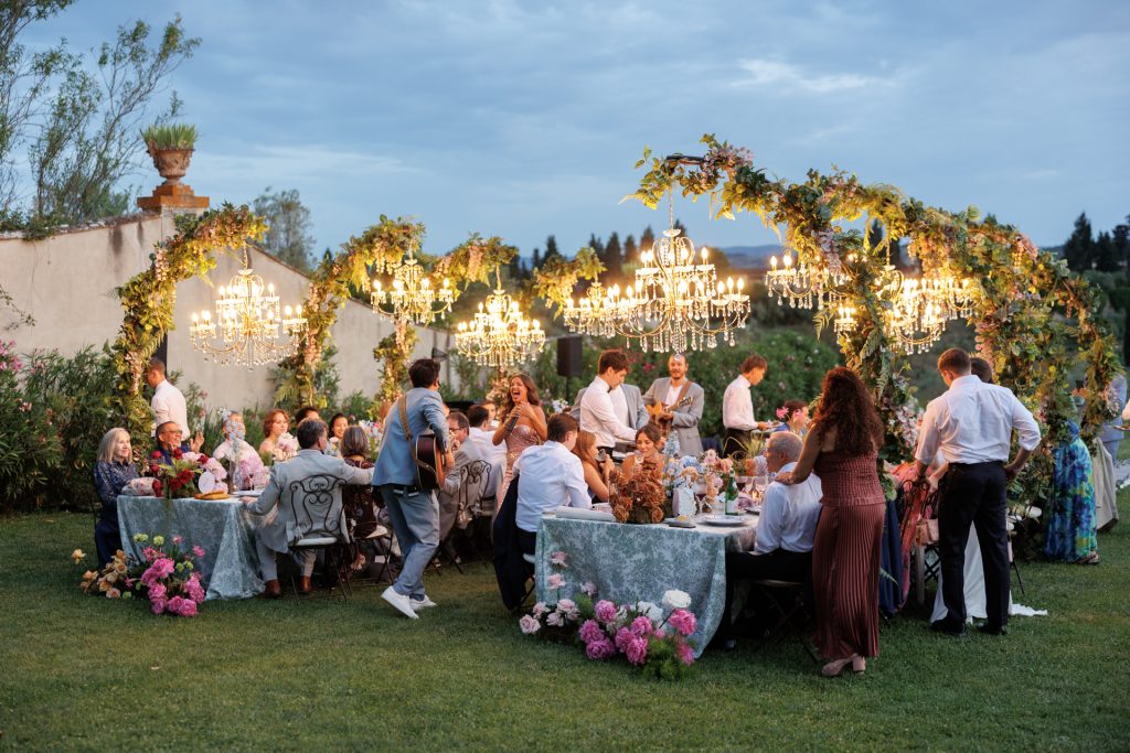 in questa foto del matrimonio di Emily e Mark firmato ester chianelli  si vede una cena elegante all’aperto in un giardino lussureggiante. Due lunghi tavoli rettangolari sono disposti parallelamente su un prato curato, coperti da tovaglie dai toni verde salvia chiaro con motivi floreali. Intorno ai tavoli sono seduti molti invitati, adulti e anziani, vestiti in abiti eleganti e colorati. Alcuni parlano, altri ridono o si godono la compagnia.

Sopra i tavoli si ergono tre grandi archi decorati con foglie verdi e fiori rosa, lilla e gialli, da cui pendono spettacolari lampadari in cristallo accesi, che diffondono una luce calda e romantica.

Un uomo con una chitarra, in abito azzurro, cammina suonando tra i tavoli, mentre una donna canta sorridendo. Il cielo è azzurro tendente al blu della sera, e sullo sfondo si intravede la campagna, un muro antico e alberi sempreverdi. Ai piedi dei tavoli, composizioni di peonie e rose color rosa, fucsia e pesca completano l’atmosfera incantata. È un momento conviviale, festoso e intimo.