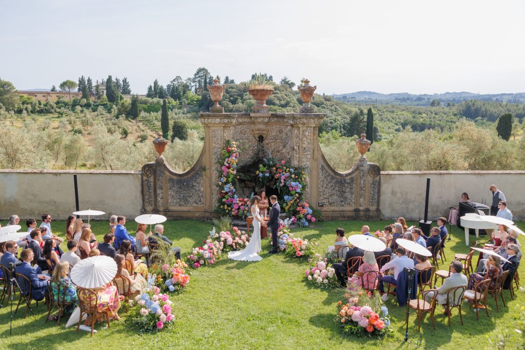 in questa foto del matrimonio di Emily e Mark firmato ester chianelli si vede un elegante giardino, gli sposi si tengono per mano al centro della scena mentre si scambiano le promesse. La sposa indossa un abito bianco lungo, scivolato e senza maniche, mentre lo sposo è in abito blu scuro. Dietro di loro, la celebrante legge da un leggio, incorniciata da una parete in pietra chiara decorata con grandi anfore in terracotta e da una fontana centrale. L’intera area è adornata da composizioni floreali rigogliose in rosa, pesca, fucsia, azzurro e arancio. Gli invitati siedono in due file ordinate di sedie in legno, alcuni con ombrellini bianchi parasole, creando un’atmosfera raffinata e romantica. Sullo sfondo si intravede un paesaggio collinare con cipressi e ulivi.