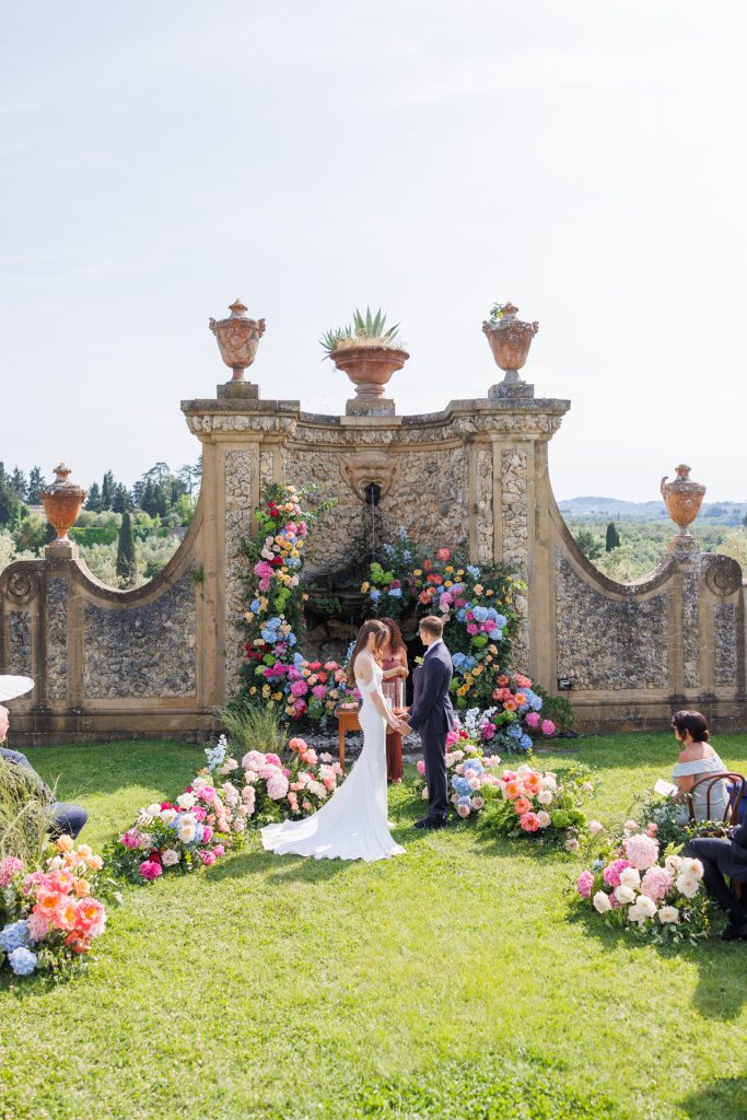 in questa foto del matrimonio di Emily e Mark firmato ester chianelli si vede La coppia di sposi è ritratta nel momento dello scambio delle promesse in un giardino elegante. La sposa indossa un abito bianco lungo e scivolato, senza maniche, mentre lo sposo è in abito blu scuro. Dietro di loro, una celebrante legge da un leggio in legno. La scena si svolge davanti a una parete in pietra chiara decorata da anfore antiche in terracotta e da una fontana con una testa scolpita. La parete è riccamente adornata da una cornice di fiori vivaci: ortensie, rose e peonie in rosa, azzurro, pesca, giallo e fucsia. Ai lati della coppia, invitati seduti osservano la scena, circondati da aiuole e composizioni floreali che creano un’atmosfera romantica e raffinata. Sullo sfondo, si intravedono colline verdi e cipressi.