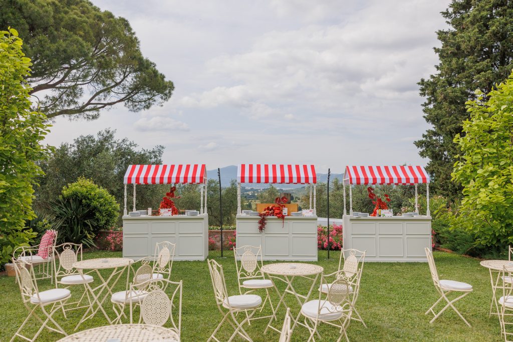 in questa foto del matrimonio di Emily e Mark firmato ester chianelli si vede  un giardino curato, circondato da alberi e cespugli verdi, si trovano tre chioschi bianchi con tetto a righe rosse e bianche. I banconi, in stile rétro, sono decorati con elementi rossi tra cui peperoncini, pomodori, fiori e bottiglie. Di fronte ai chioschi ci sono tavolini e sedie in ferro battuto bianco con cuscini bianchi, disposti ordinatamente sul prato. Sullo sfondo, si intravede un paesaggio collinare con vista panoramica, sotto un cielo parzialmente nuvoloso. L’atmosfera è rilassata, elegante e festosa, ideale per un ricevimento all’aperto.