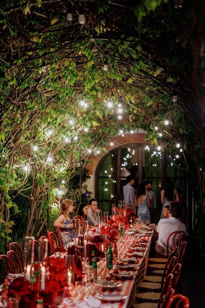 in questa foto del matrimonio di Emily e Mark firmato ester chianelli si vede la scena di una Cena serale sotto le luci in giardino. Tavola apparecchiata in stile italiano, ospiti seduti, chiacchiere e atmosfera rilassata. La pergola è illuminata da tante lucine sospese tra i rami. Il colore rosso dei fiori e delle sedie contrasta con la luce calda e soffusa della sera.