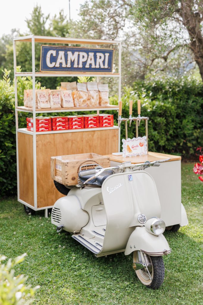 in questa foto del matrimonio di Emily e Mark firmato ester chianelli si vede Corner Campari con Vespa vintage. Un elegante chiosco all’aperto con insegna “CAMPARI” in stile retrò. Sotto la struttura in legno ci sono snack e bottigliette rosse. Di fronte, una Vespa color panna con una cassetta di legno sul retro funge da punto di appoggio per il bar.