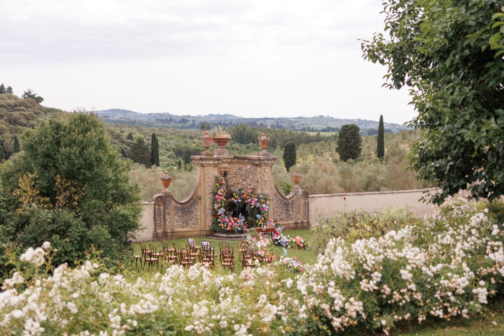 in questa foto del matrimonio di Emily e Mark firmato ester chianelli si vede un giardino fiorito e rigoglioso, davanti a un’elegante parete in pietra decorata con vasi in terracotta e una nicchia centrale, sono disposte in semicerchio alcune sedie in legno scuro con schienale curvo. La parete, in stile classico, è adornata da una cascata di fiori colorati: rose, ortensie e peonie nelle tonalità del rosa, arancio, blu e lilla. Intorno, siepi di rose bianche incorniciano la scena. Sullo sfondo, si estende un paesaggio collinare verdeggiante, punteggiato da cipressi e ulivi, tipico della campagna toscana. L’atmosfera è romantica, tranquilla e immersa nella natura.