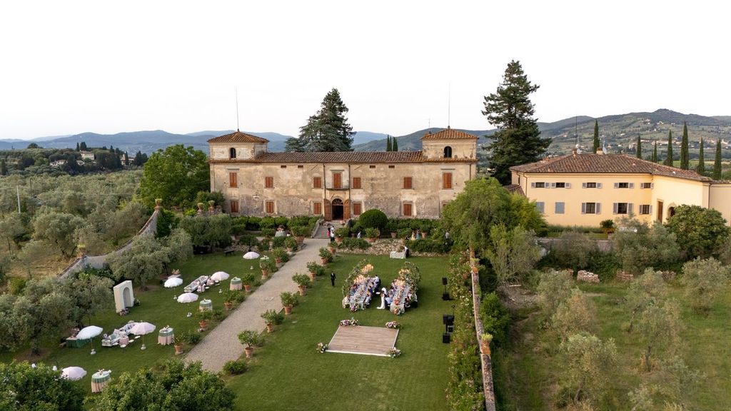 in questa foto del matrimonio di Emily e Mark firmato ester chianelli si vede una vista aerea panoramica di una villa storica in campagna, circondata da colline di colore verdi, oliveti e cipressi tipici del paesaggio toscano. L’edificio principale è una villa antica in pietra dai colori ocra e rosati, con tetti in cotto e finestre con persiane in legno. Accanto, si estende una struttura più moderna e chiara, sempre integrata nel contesto.

Nel giardino centrale, ordinato e curato, si sta svolgendo un ricevimento all’aperto. Lungo un viale di ghiaia ci sono tavolini tondi decorati con tovaglie rosa e verde salvia, sedute in ferro battuto e ombrelloni di colore bianchi e rosa con frange vintage. Più avanti, sulla destra, si vedono due lunghe tavolate parallele per la cena, ornate con fiori e candelabri, circondate da invitati seduti.

In fondo al giardino è stato posizionato un palco in legno per l’intrattenimento serale, mentre l’intero evento è immerso in un’atmosfera elegante, romantica e immersa nella natura.