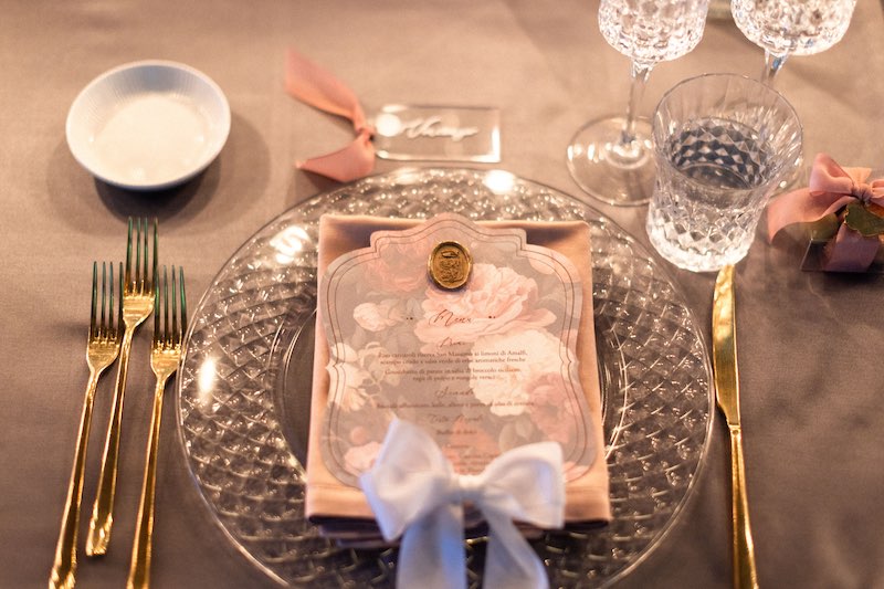 In questa foto, un dettaglio della mise en place del matrimonio di Maria Pia e PierPaolo con un sottopiatto trasparente, posate d'argento, bicchieri in vetro trasparente, tovagliolo rosa, menù con stampa floreale e un nastro di colore bianco